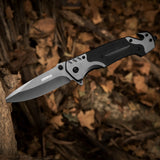 A folding pocket knife with a G10 handle, silver blade, and black accents, featuring a pocket clip, placed on a wooden surface with leaves in the background.
