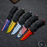 Five folding pocket knives with black handles are arranged diagonally on a dark gray slate surface. Each knife has a different colorful blade design, including blue, yellow, light blue, red, and orange patterns.