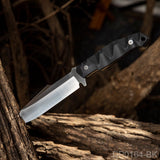 A knife with a black G10 handle and a partially serrated blade, resting on a wooden branch.